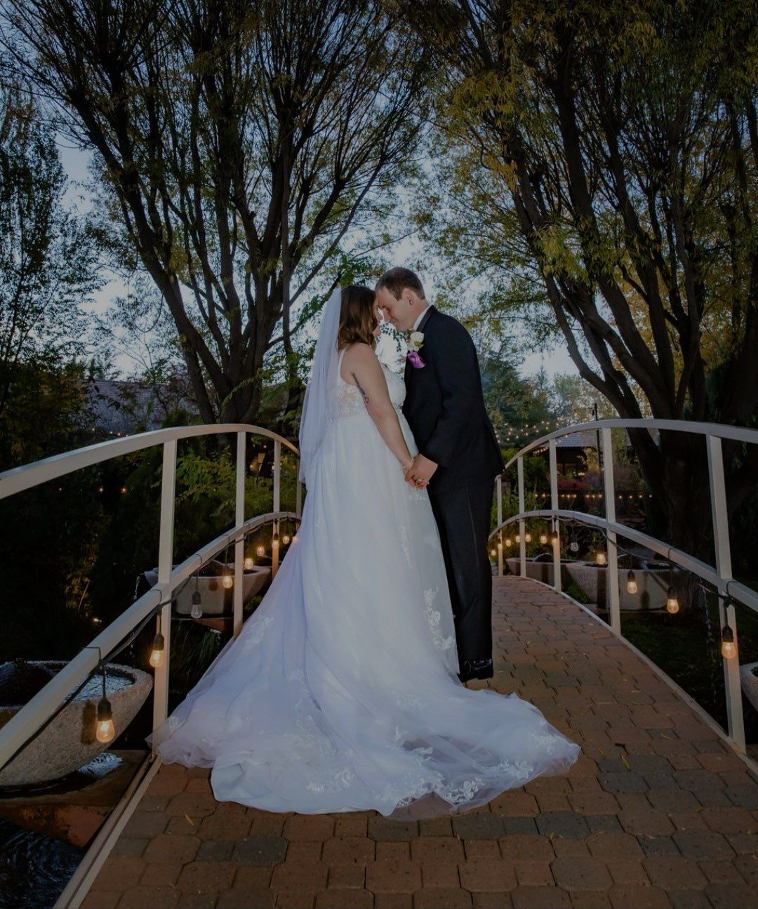 Bride and groom hold hands on a bridge, posing for a photo. Evening setting with string lights and trees.