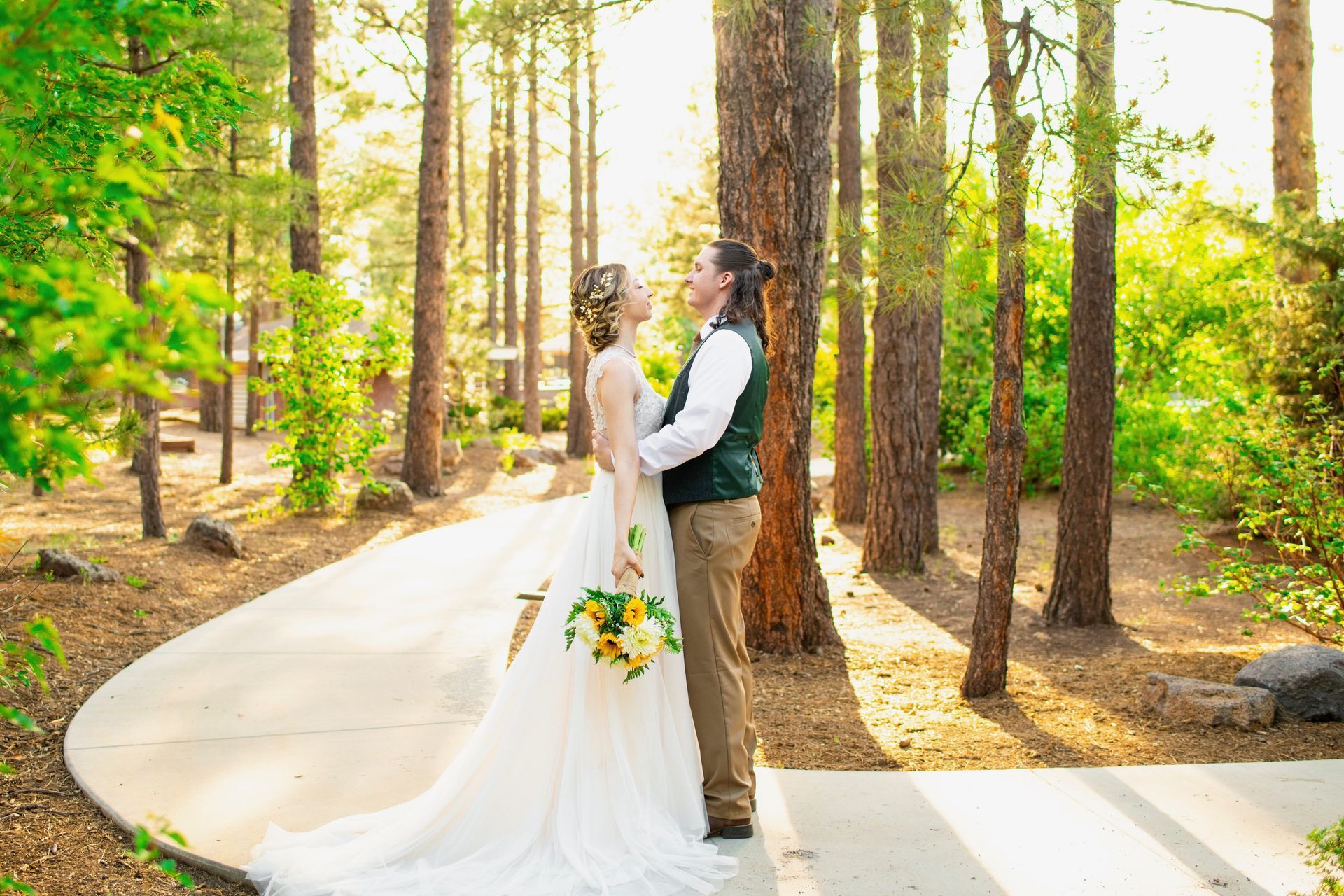 Couple embraces on a path in a sunlit forest, bride in a white gown, groom in vest.