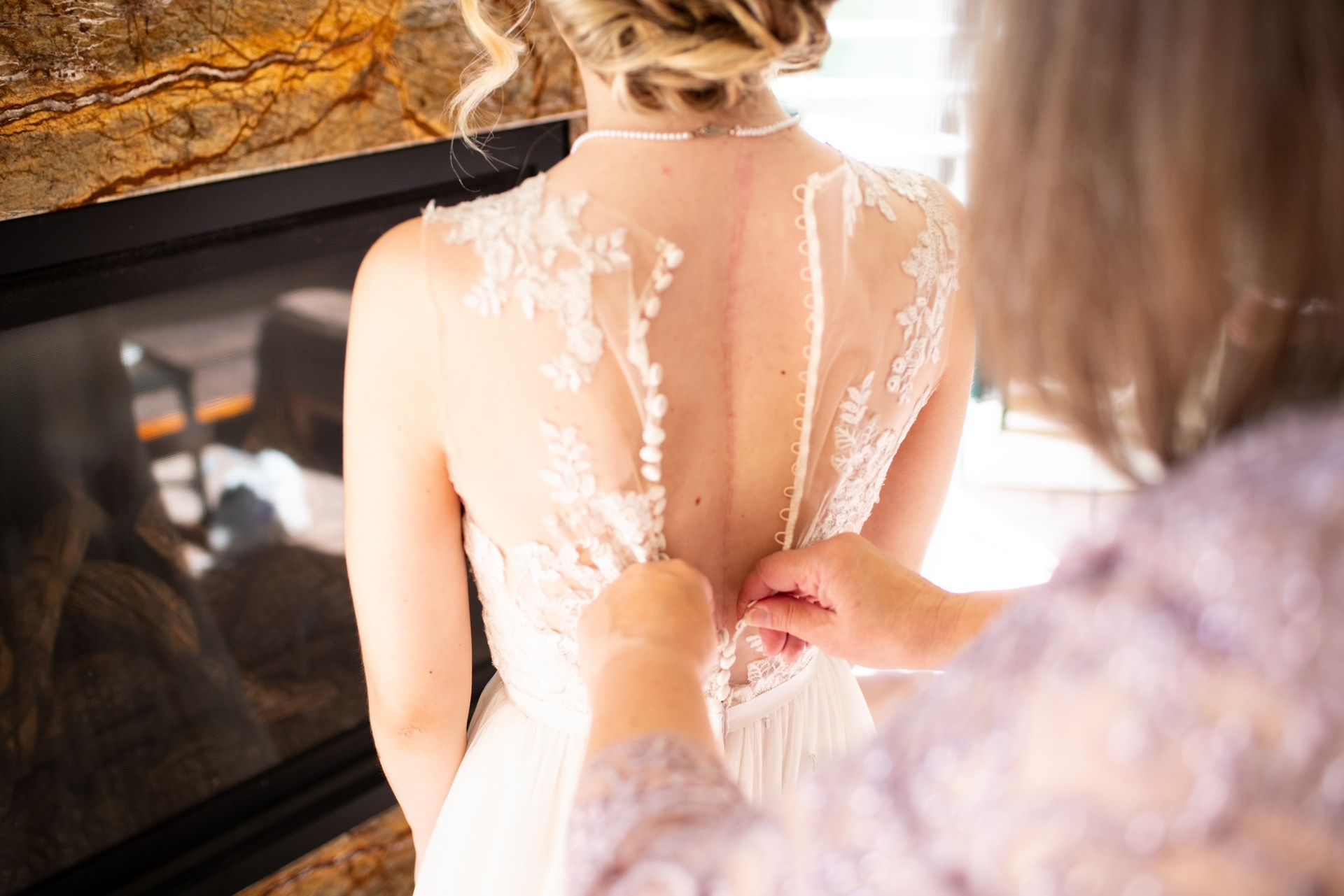 A person's wedding dress being buttoned up by another person; near a fireplace.