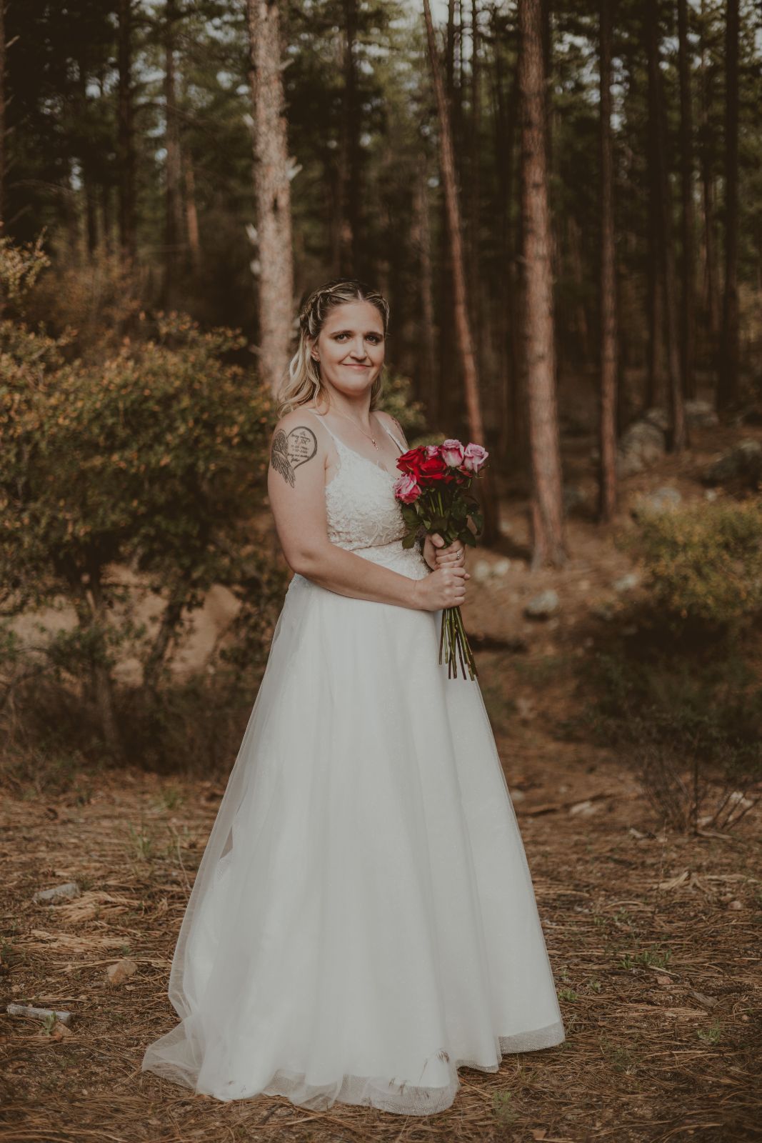 Bride in white wedding dress holding flowers, standing in a forest.