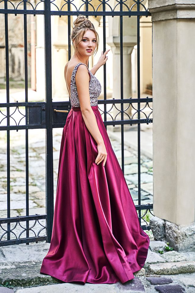 Woman in a maroon satin gown, posing by a black gate, looking over her shoulder.