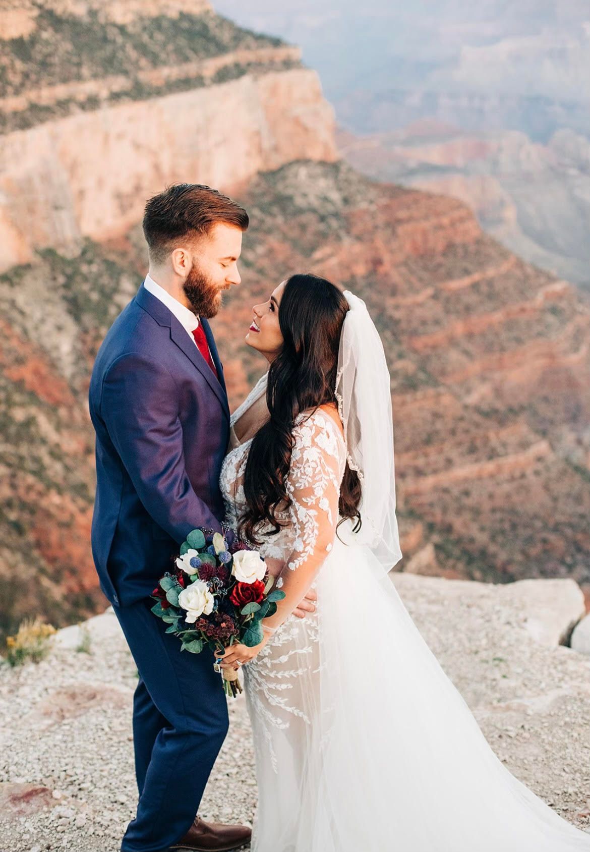 Couple in wedding attire at Grand Canyon, smiling and embracing, bouquet in hand, scenic background.