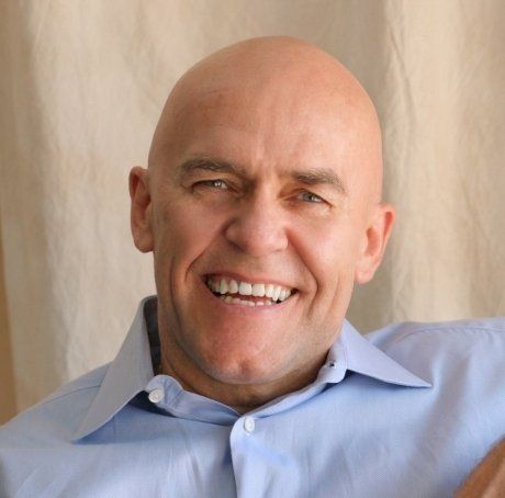 Bald man wearing a blue button-down shirt smiles at the camera. White backdrop.