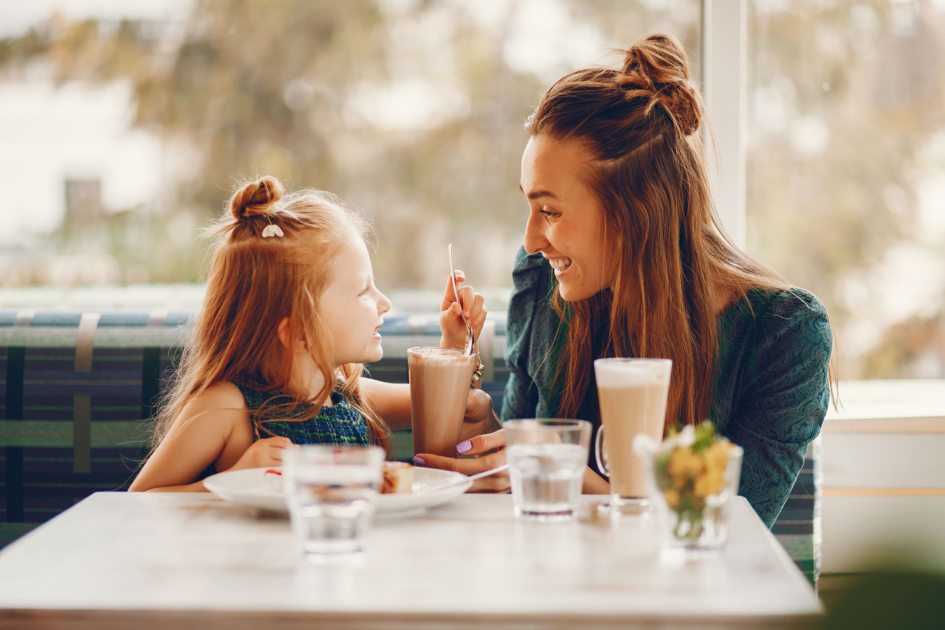 A woman and young girl with similar hairstyles share smiles over milkshakes at a cozy cafe.