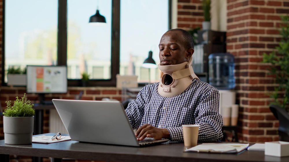 Man wearing neck brace, typing on laptop in office.