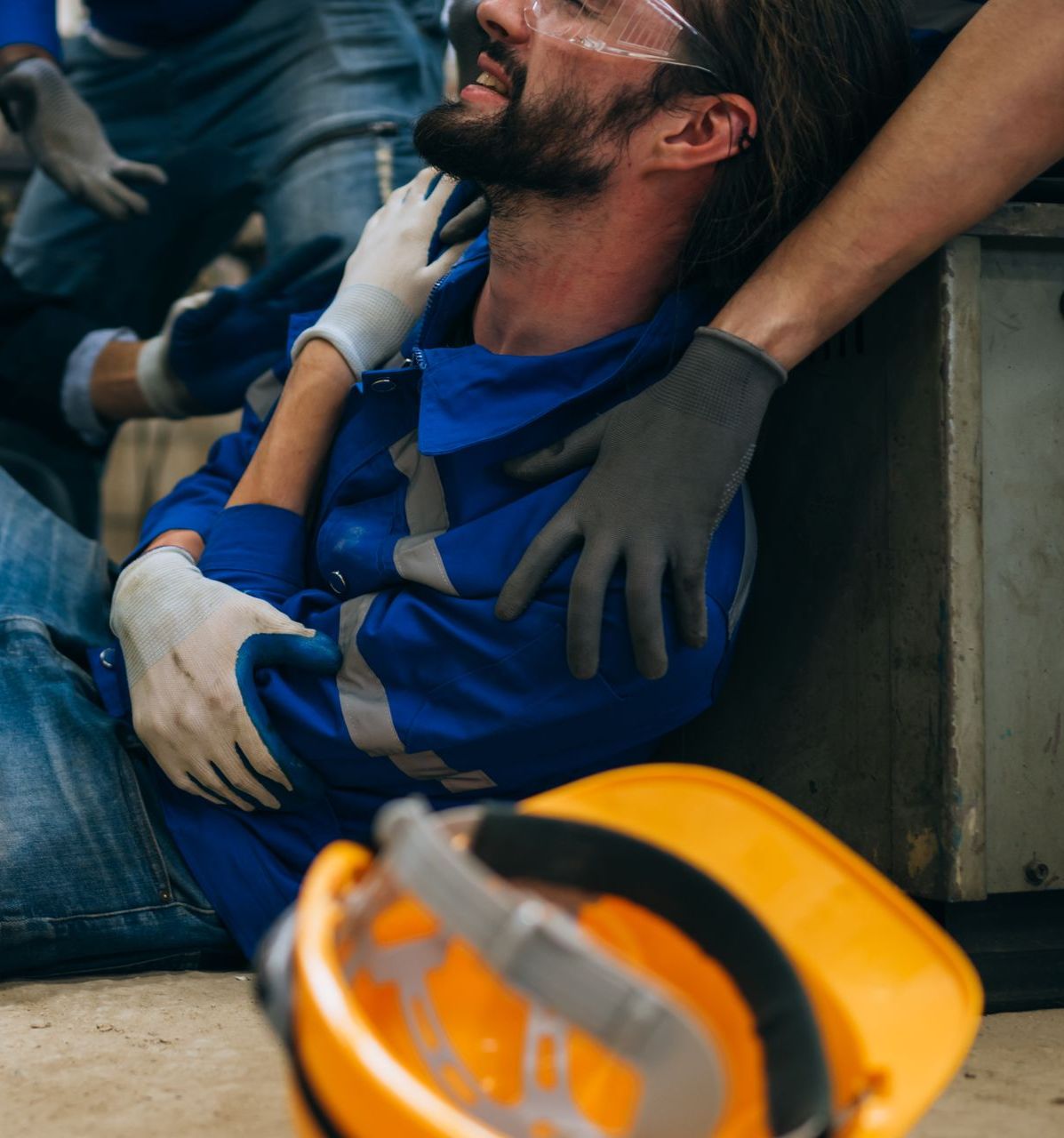 Man in blue work suit slumped on floor, being assisted by others; safety helmet nearby.