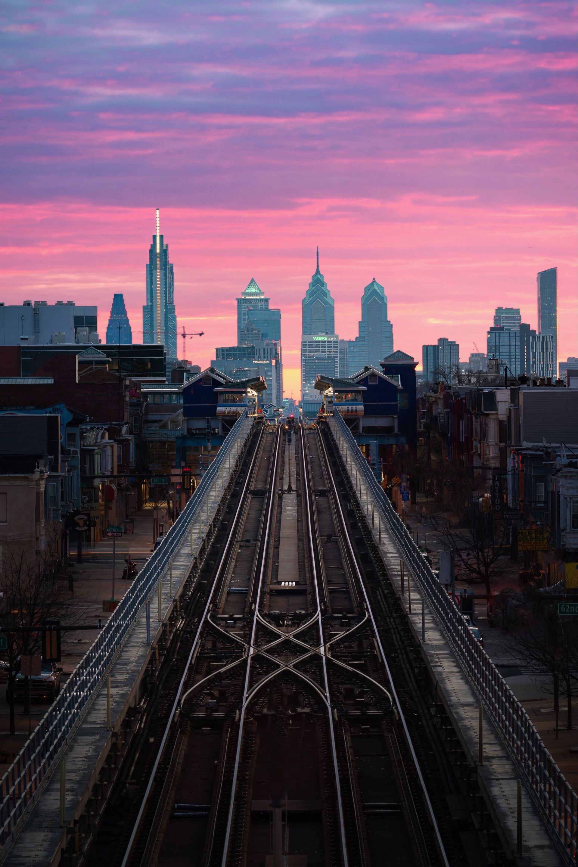 Philadelphia skyline at sunset, seen from elevated train tracks under vibrant pink and purple sky.