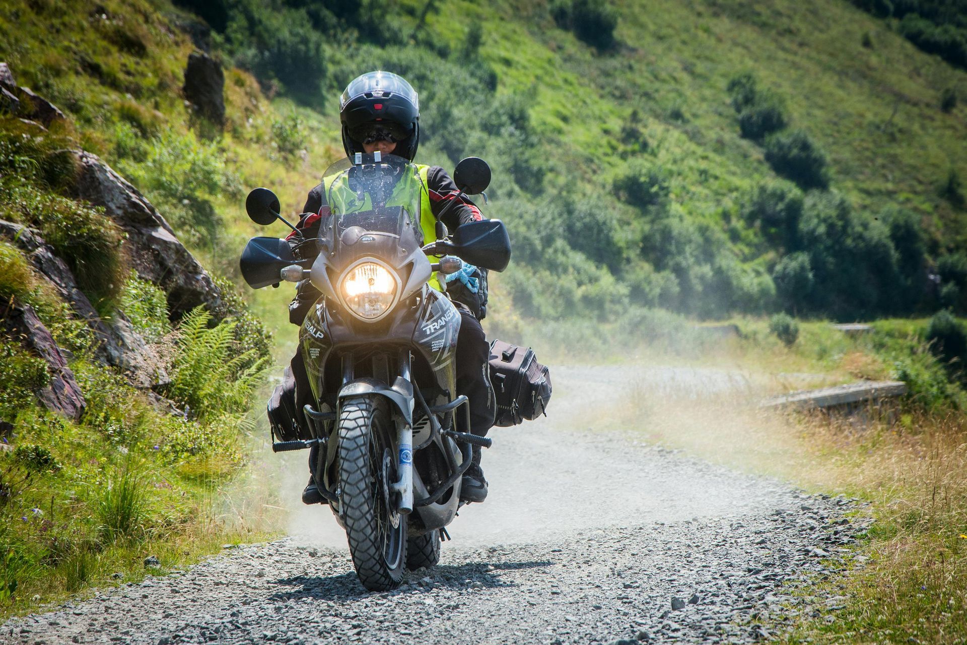 Motorcyclist on dirt road in mountains; green vegetation and rocks line the way.