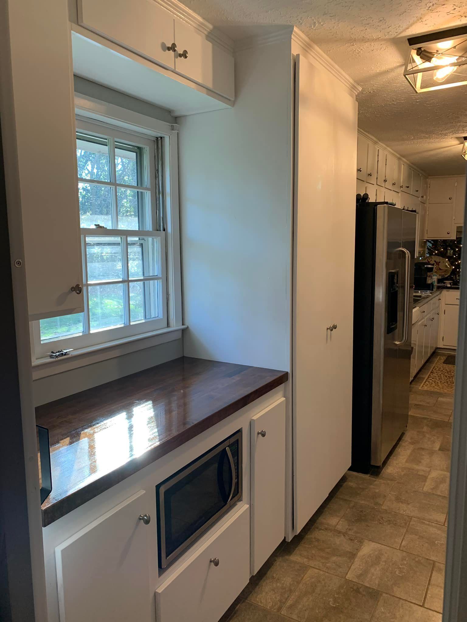 A kitchen with white cabinets , a microwave , a refrigerator and a window.