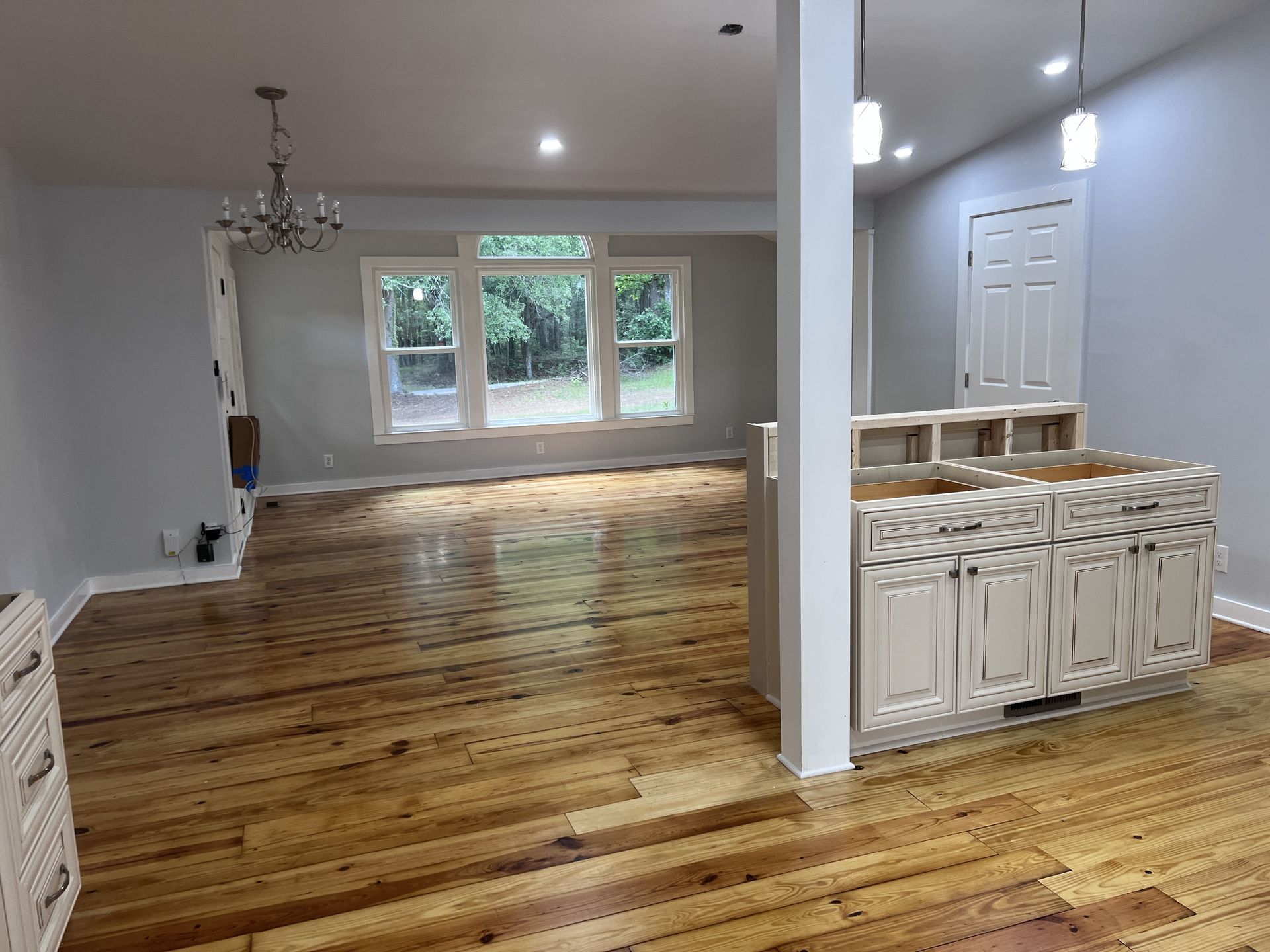 Empty renovated living space with hardwood floors and light-colored walls, kitchen cabinets on the right.