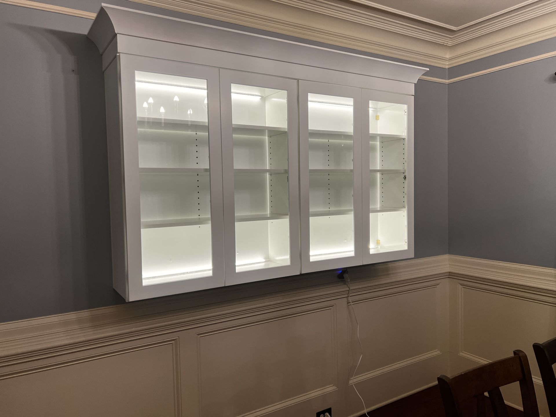 White wall-mounted cabinet with glass doors, lit shelves against a gray wall.