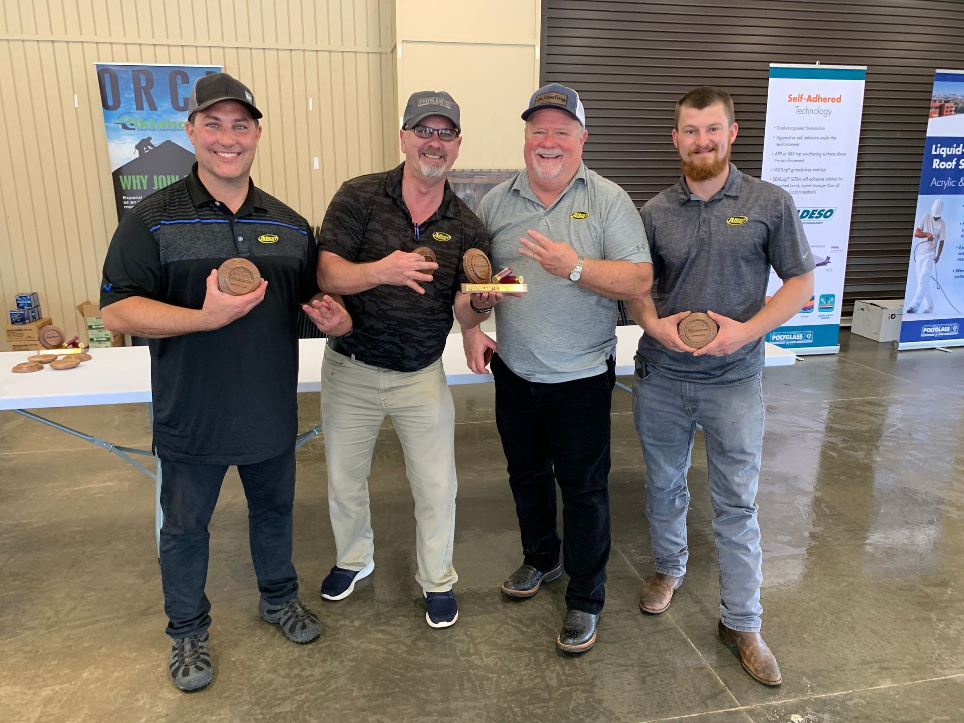 Four men holding small trophies, posing indoors with signs in the background.