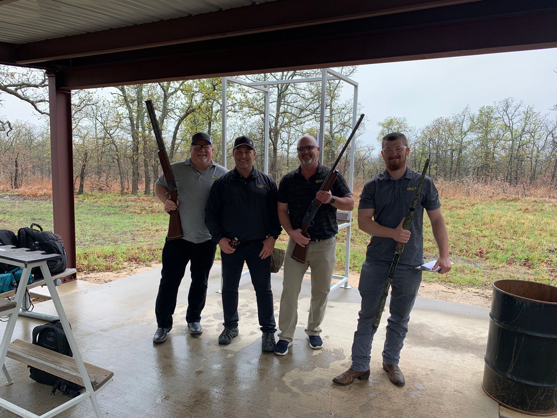 Four men with rifles under a shelter. They stand on a concrete surface, in front of a wooded area.