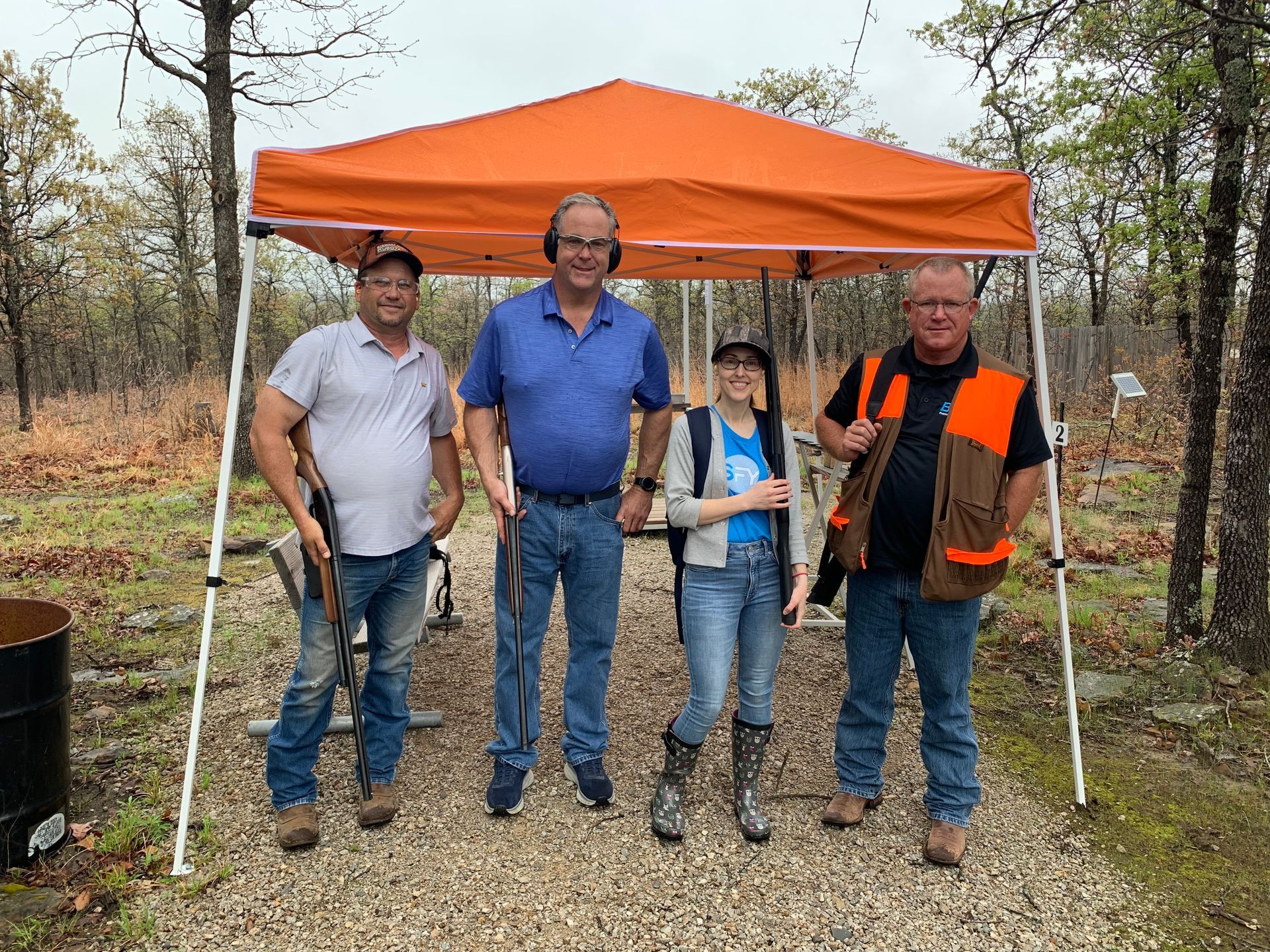 Four people stand under an orange canopy at an outdoor shooting range. Two hold firearms.