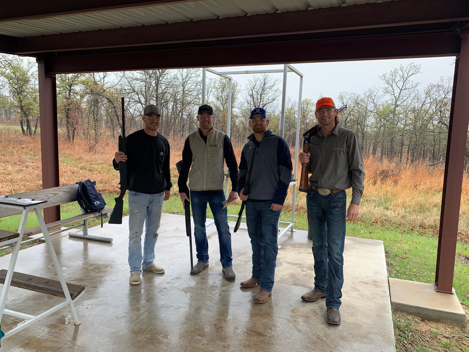 Four people stand holding rifles at an outdoor shooting range under a covered area.