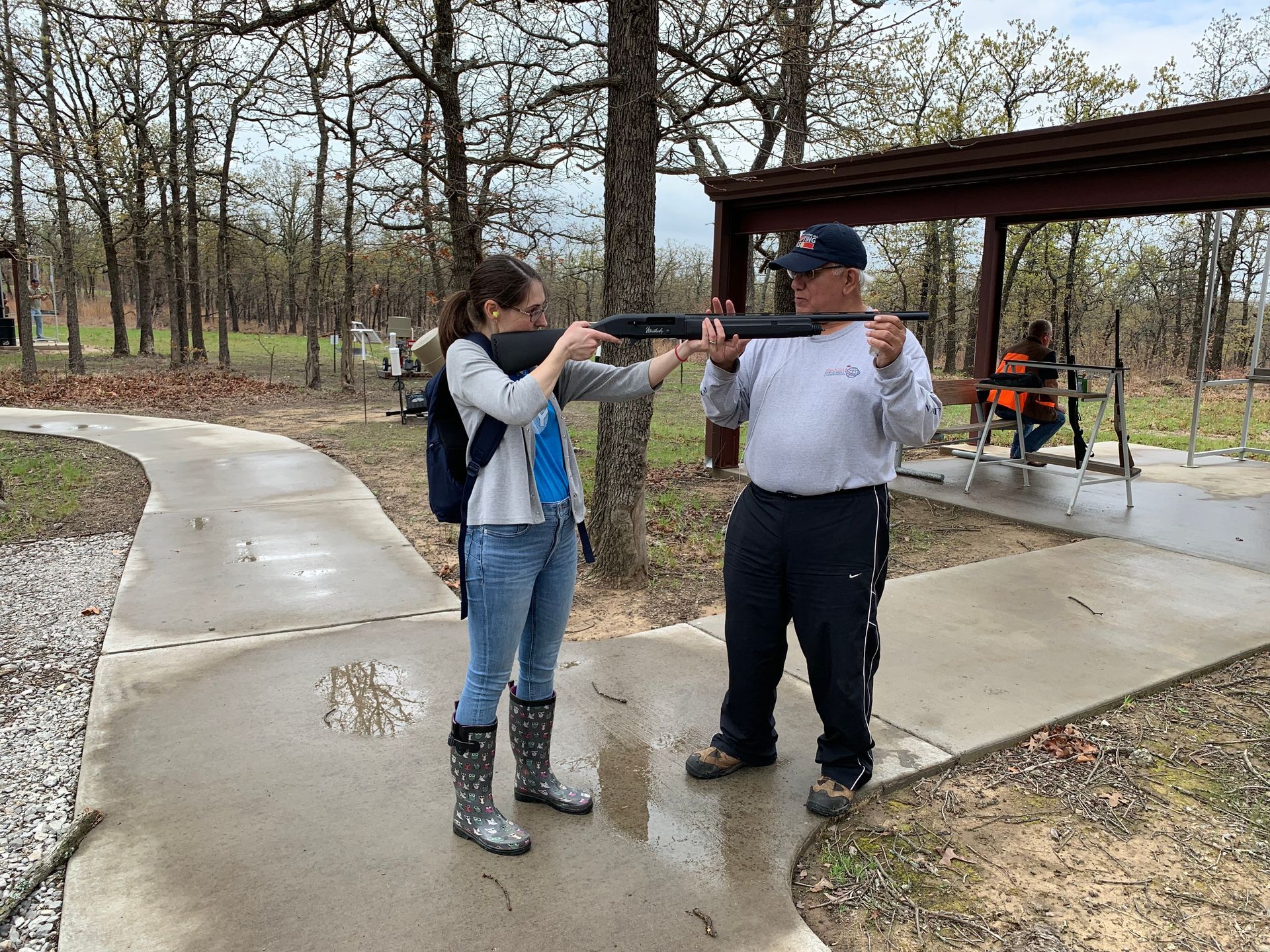 Person aiming firearm with guidance from another person outdoors. Concrete path, trees visible.