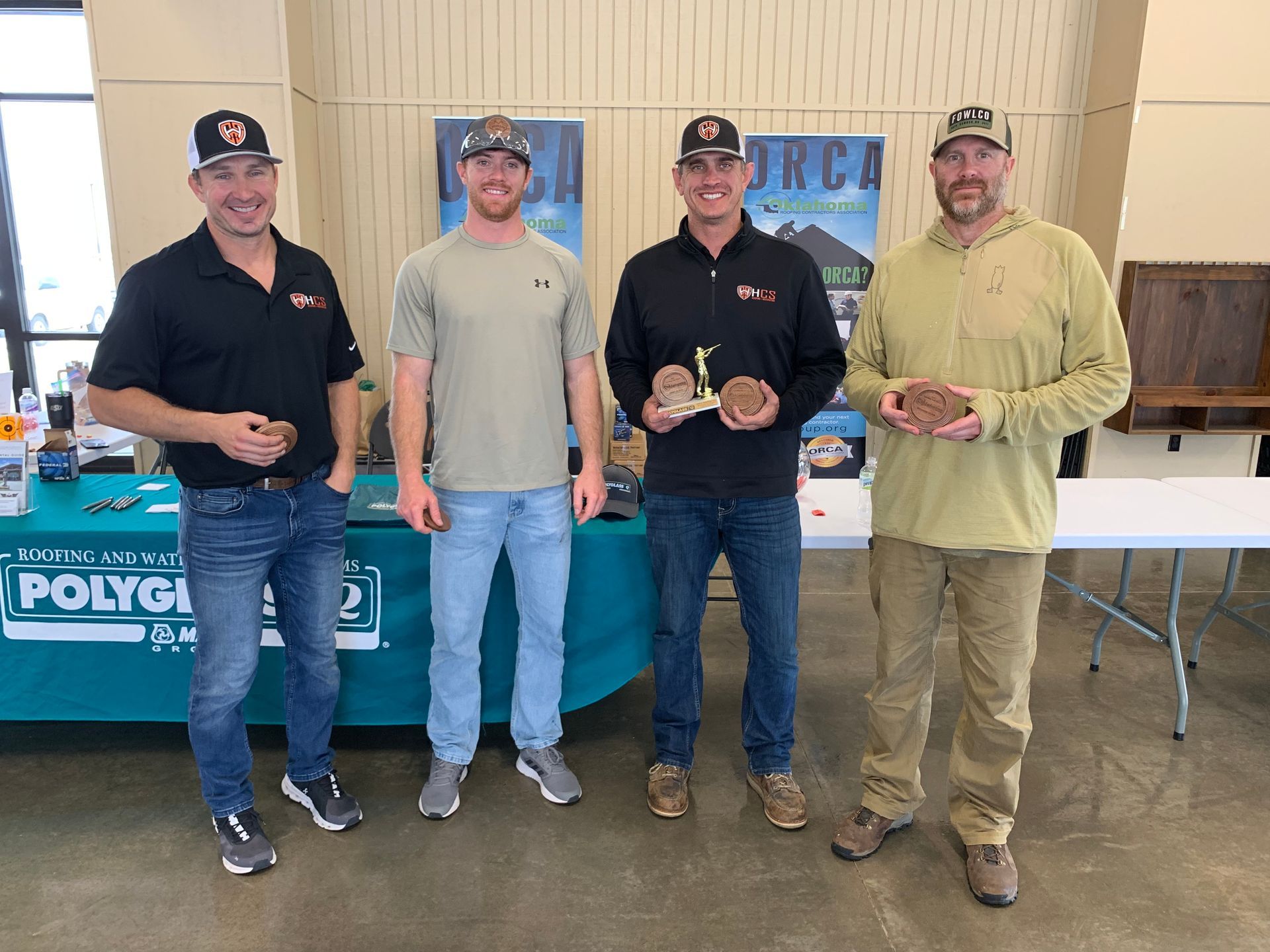 Four men in caps stand with prizes at an event table, holding round, brown objects.