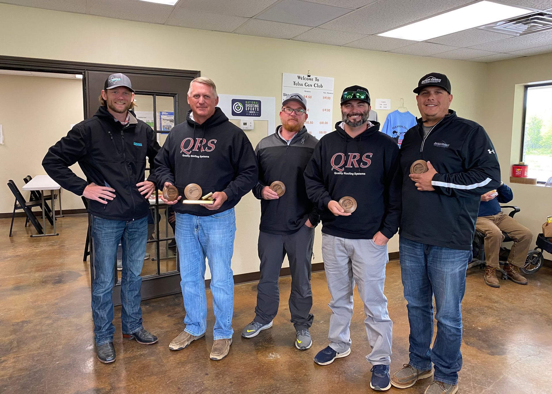 Five men holding trophies, posing in a room, some wearing hoodies with 