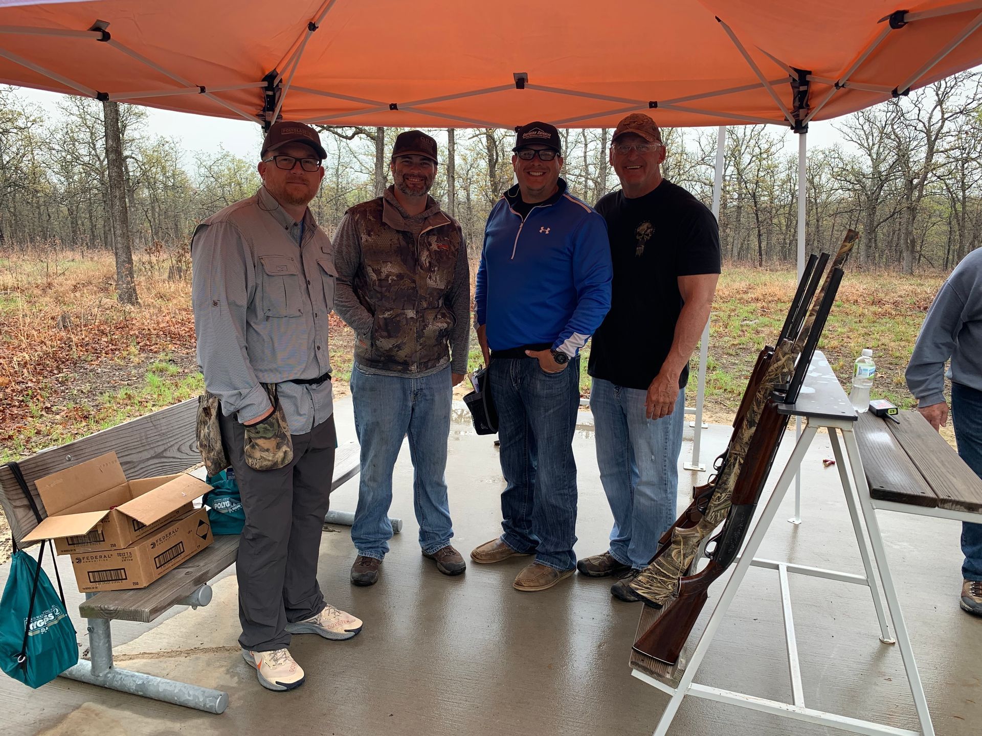 Four men under a canopy with shotguns, standing at a shooting range.