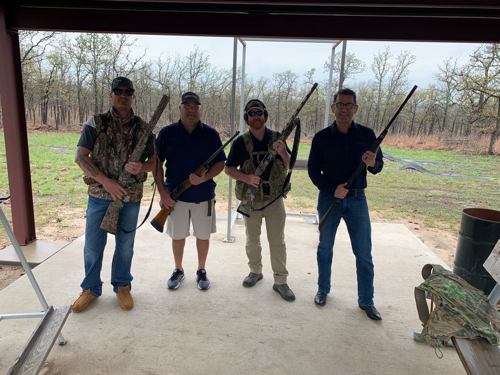 Four people with shotguns at an outdoor shooting range. They are standing under a covered area, and the ground around them is a mix of concrete and grass.