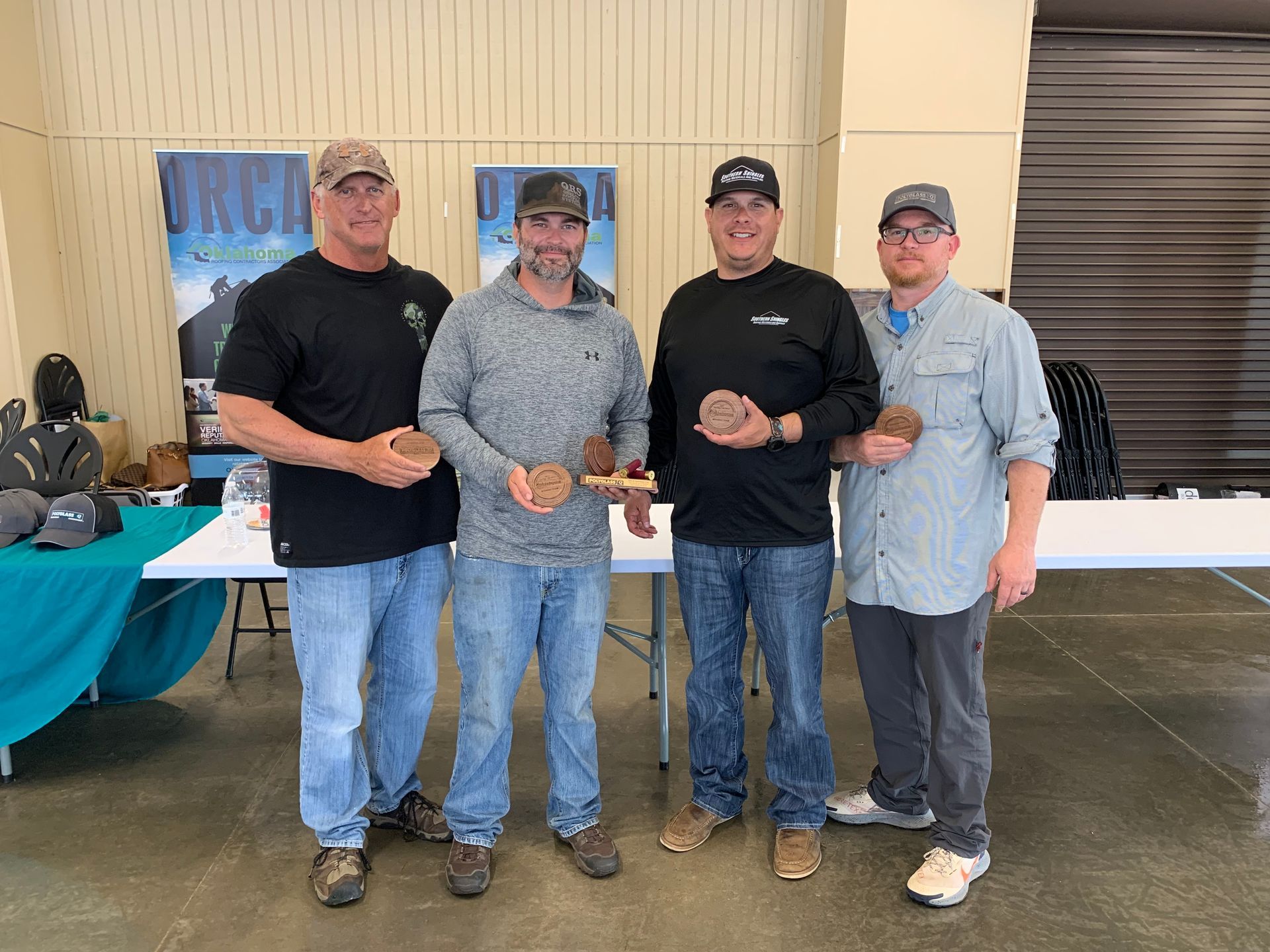 Four men hold brown objects, possibly trophies, at an event with a display table.