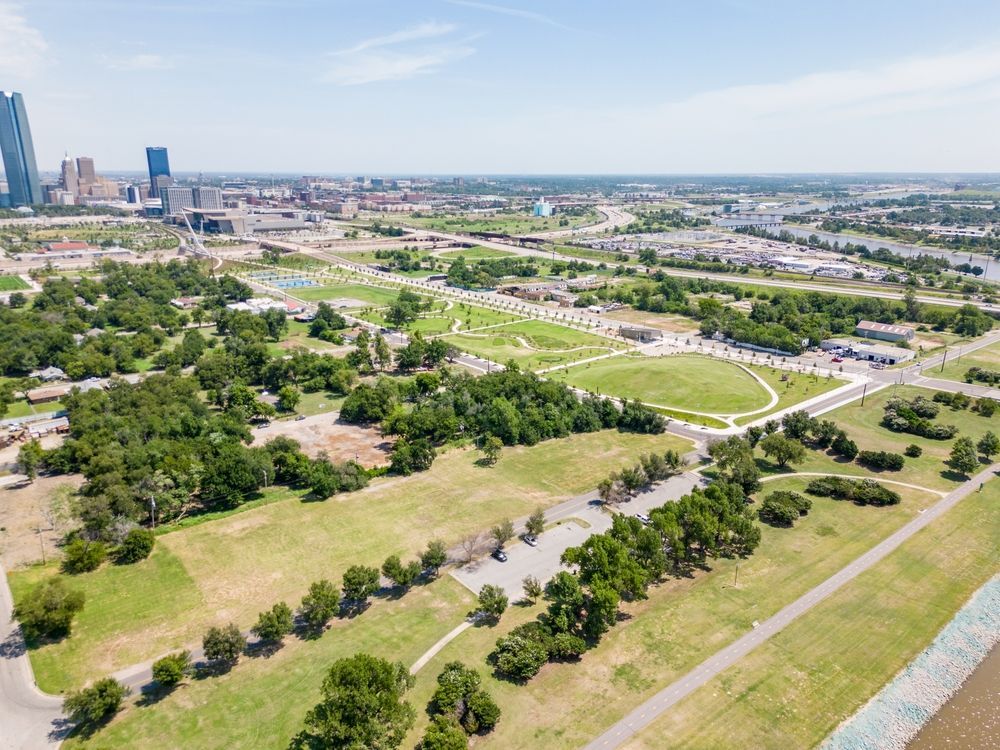 Aerial view of a park with green fields and trees, city skyline in the background, blue sky overhead.