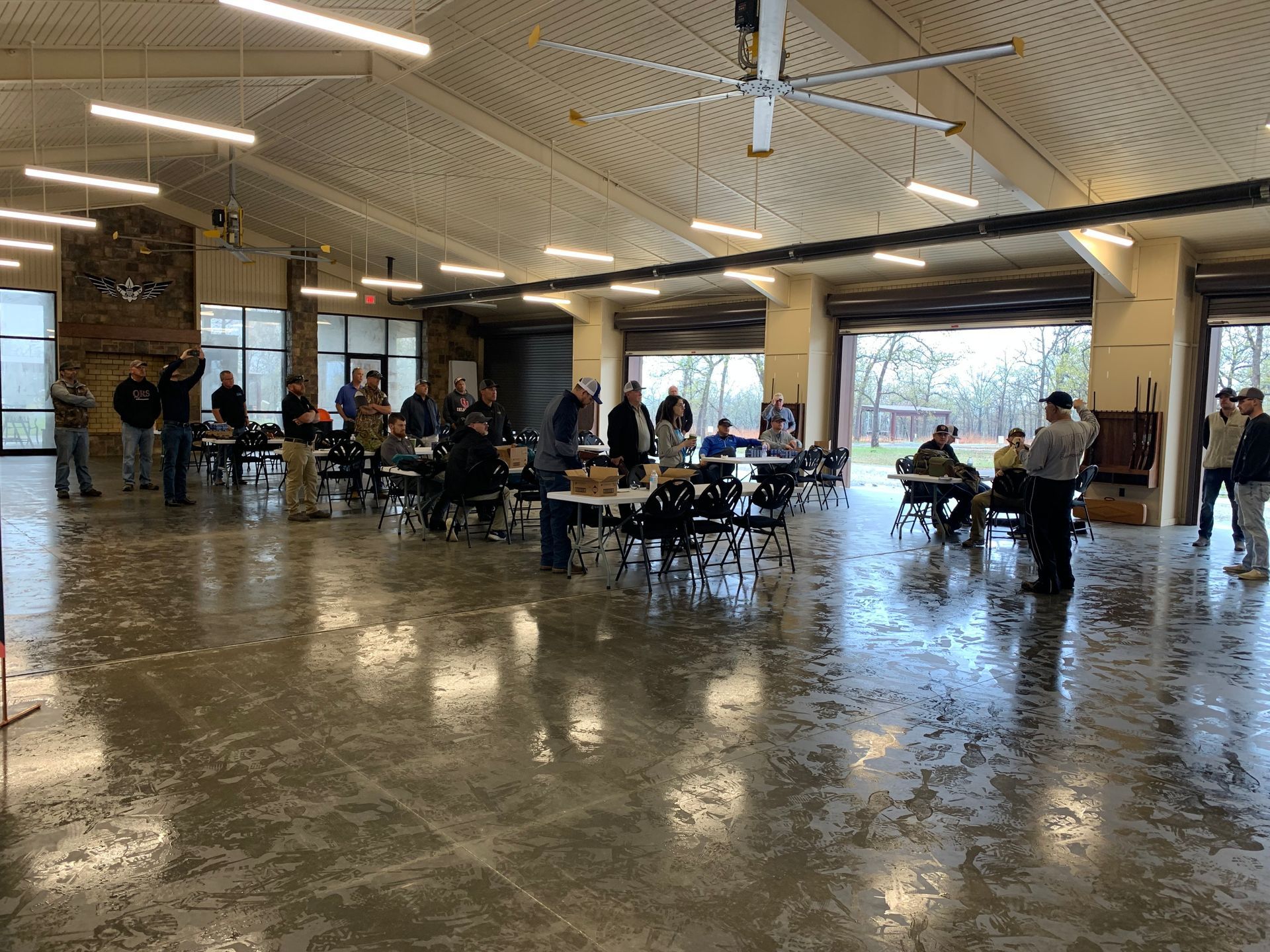 Large indoor gathering: people at tables, some standing. Concrete floor, high ceiling, windows, and open doors.