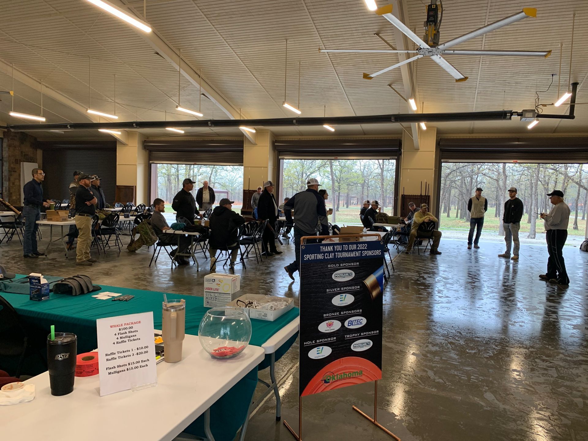 People at an event inside a large building with roll-up doors open to a wooded area. Sign at front.