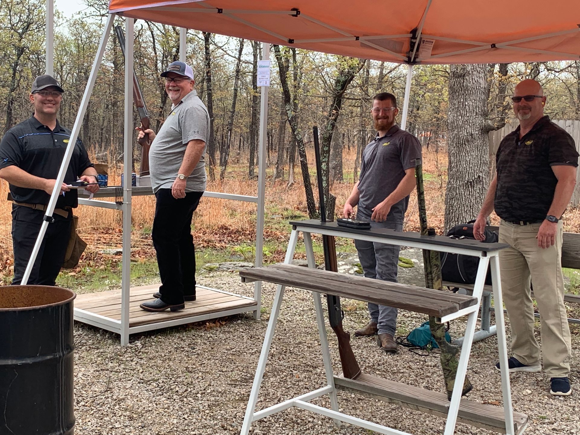 Four men at an outdoor shooting range under a canopy. Guns are on a table.