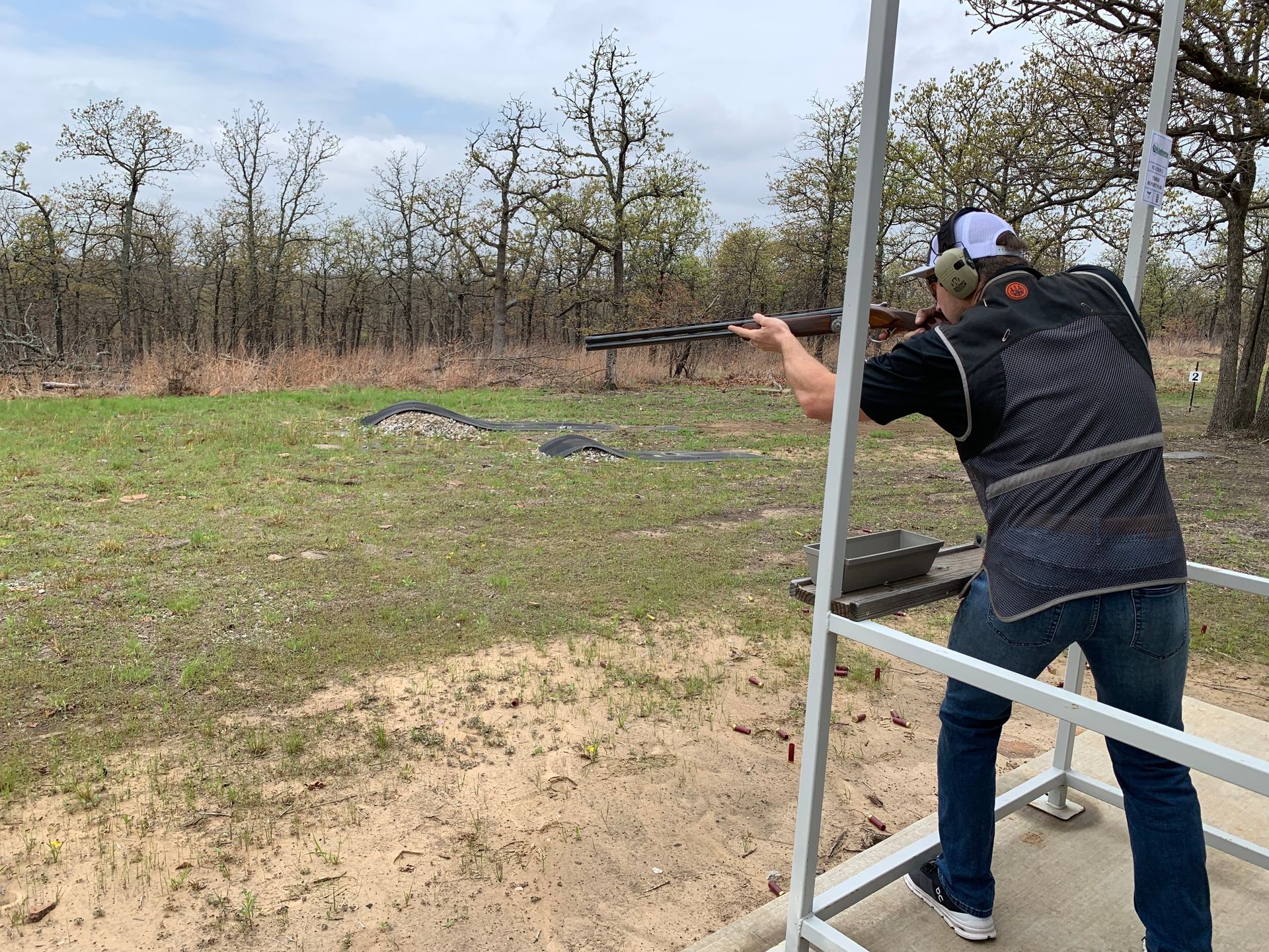 Person shooting a shotgun at a clay target on a shooting range, wearing a vest and hat.