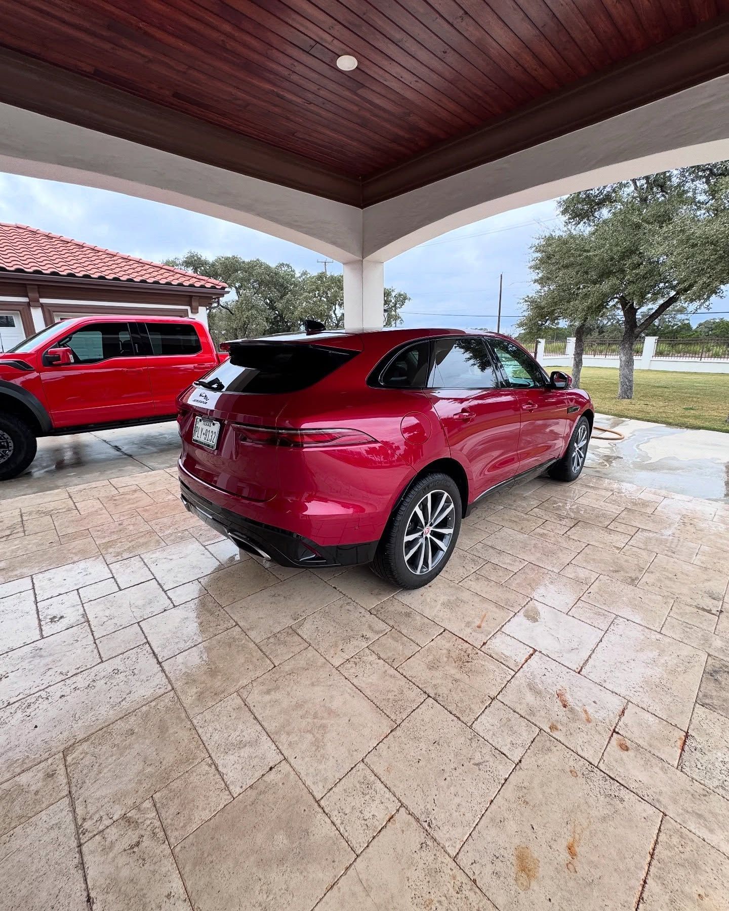 Red Jaguar SUV parked under a covered structure with a red truck in the background.