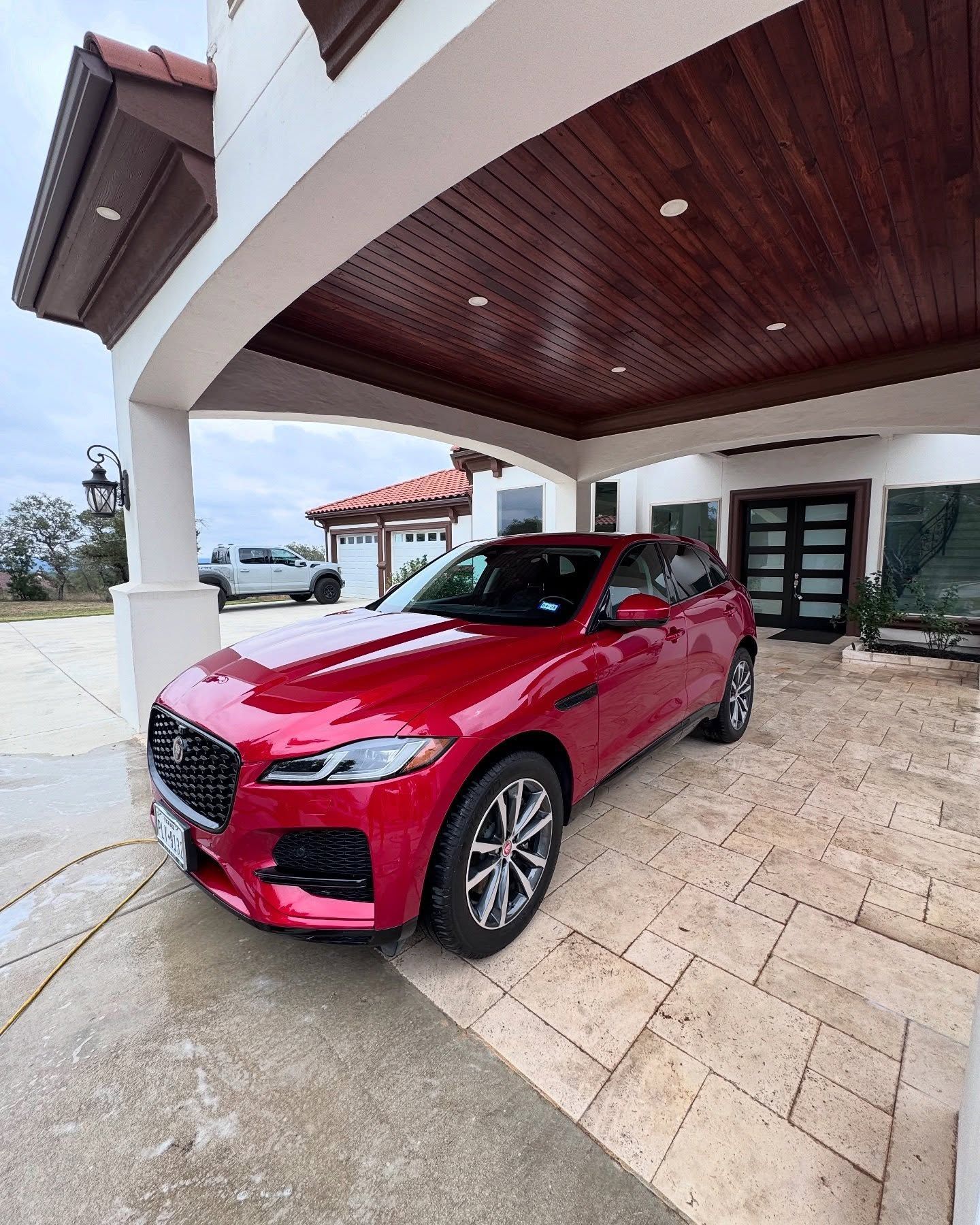 Red Jaguar SUV parked under a home's covered entrance with a light-colored brick floor and dark wood ceiling.