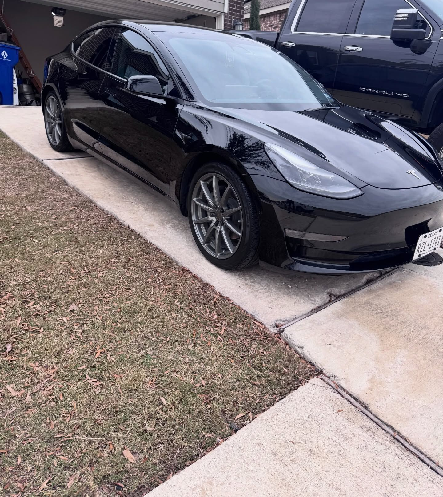 Black Tesla Model 3 parked on a driveway beside a dark pickup truck.
