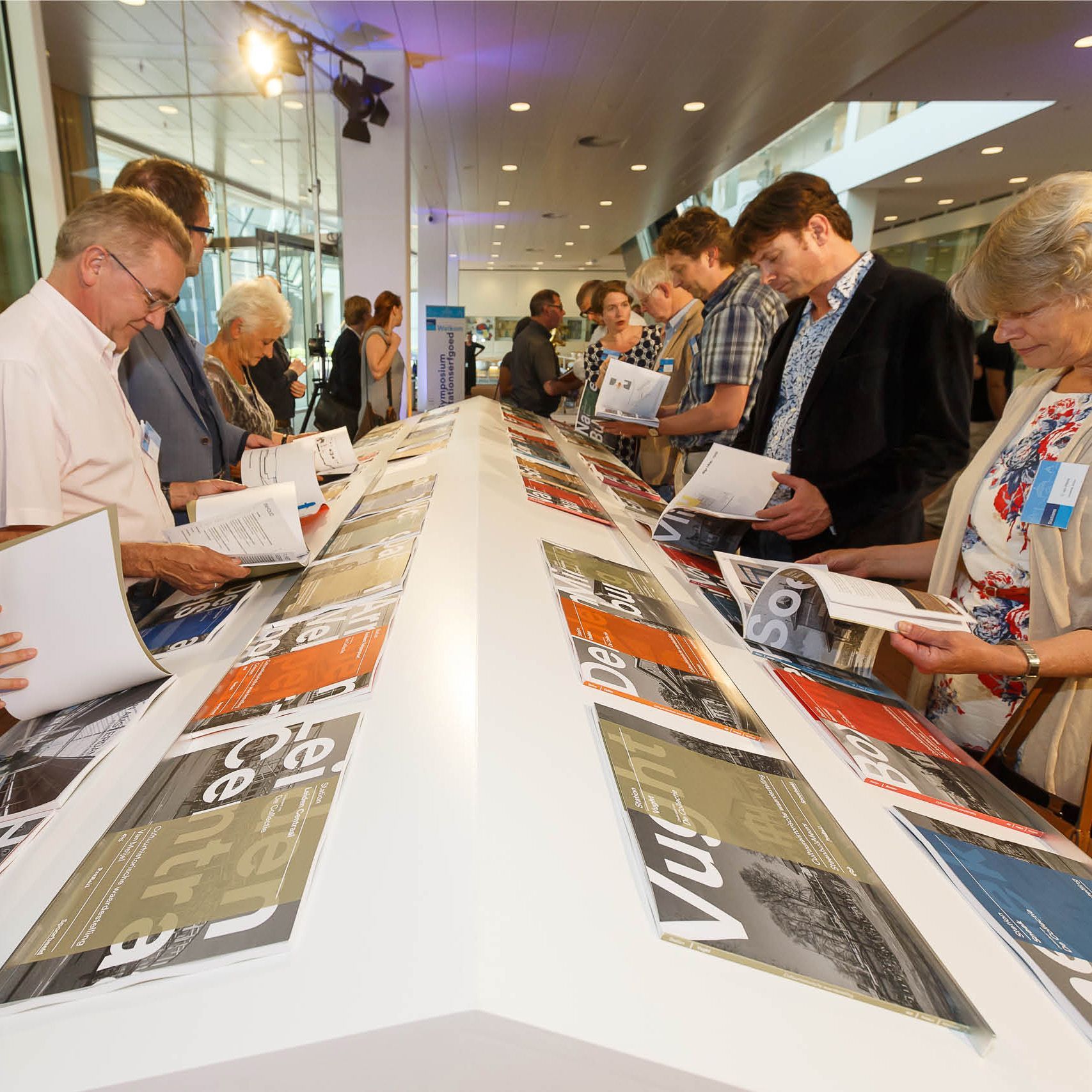 Mensen bekijken de tentoongestelde voorwerpen op een lange witte tafel binnenshuis, met licht van boven.
