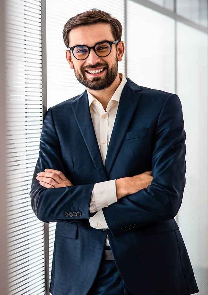 Man in a navy suit and glasses, smiling with arms crossed, leaning against a window in a modern office.