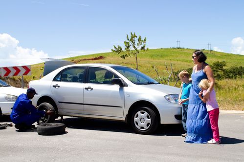 Man changing a tire on a silver car while a woman and two children watch on a sunny roadside.