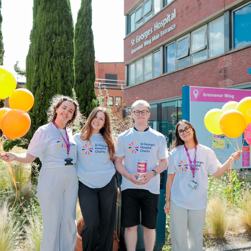 Four people wearing matching charity shirts pose with yellow and orange balloons outside St George's Hospital.