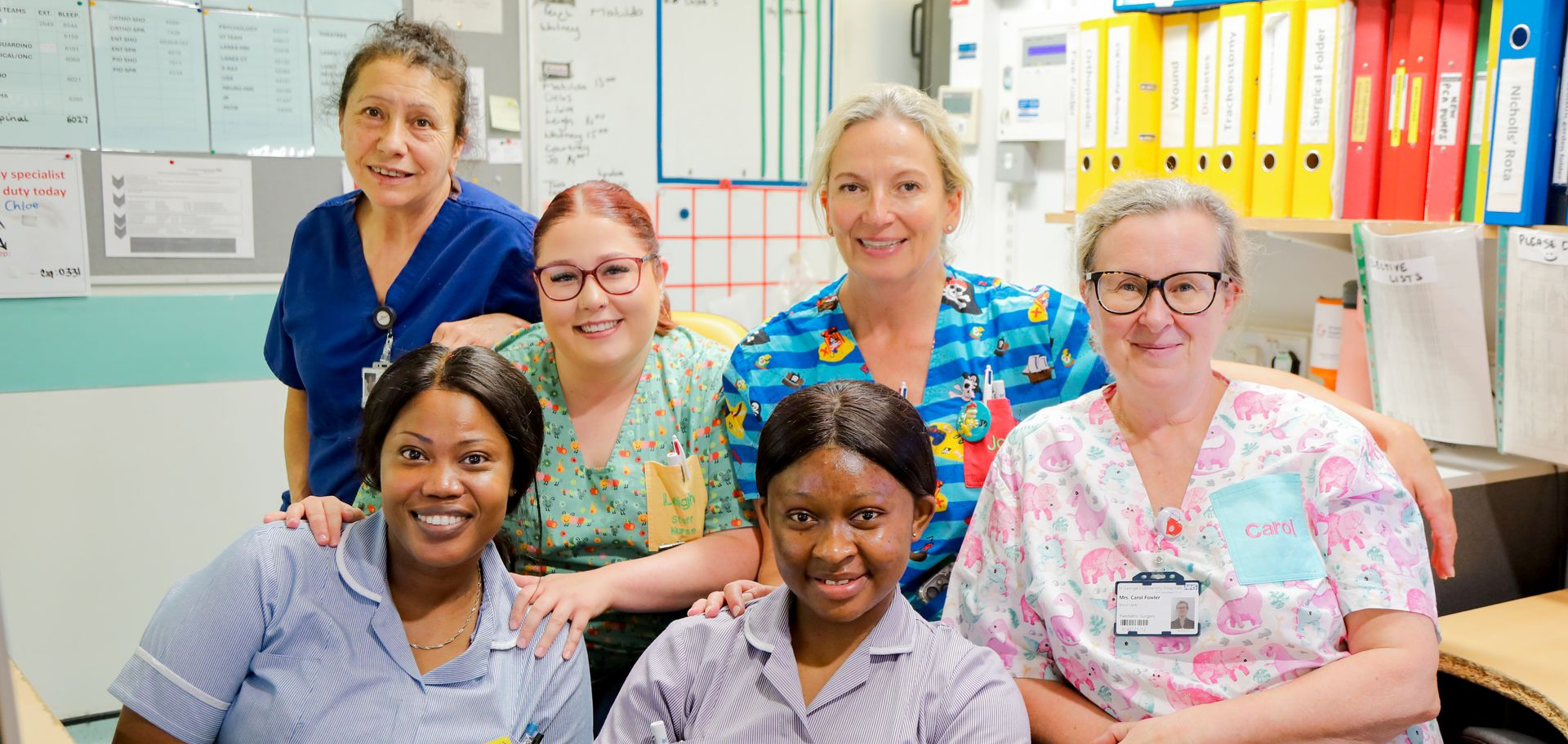 A group of six smiling healthcare staff in scrubs and uniforms standing together in a brightly lit office space.