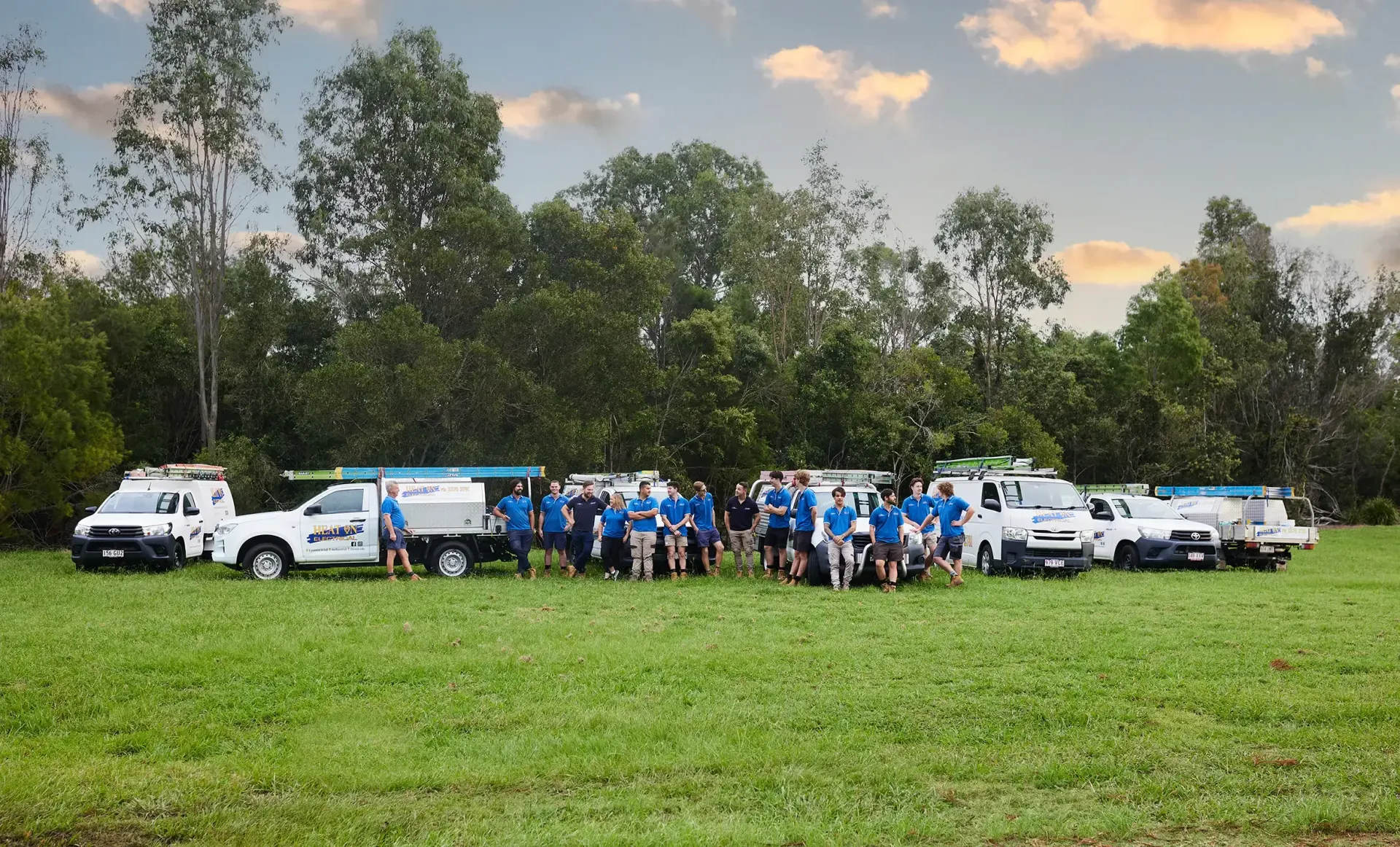 Group of people in blue shirts posing with white trucks on a grassy field at sunset