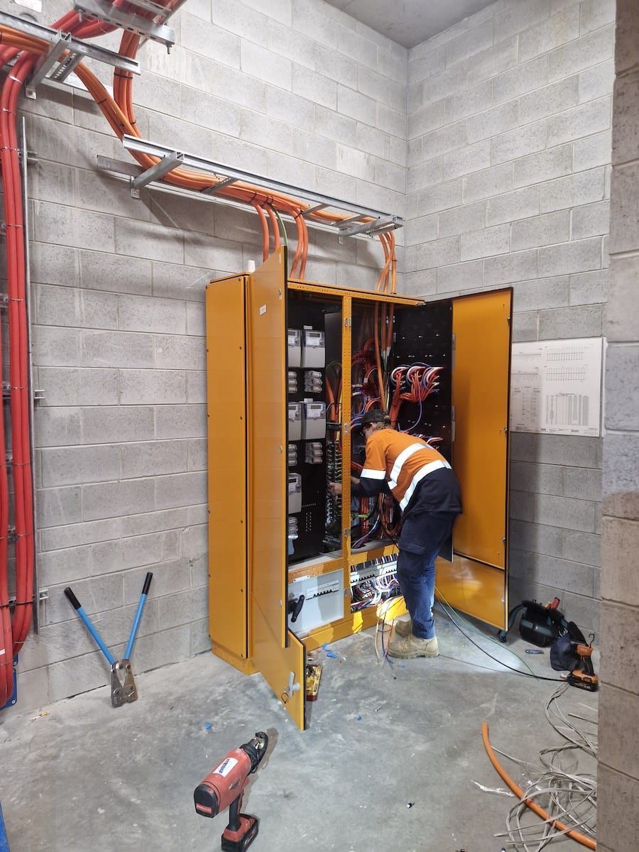 Worker inspecting electrical cabinet in a concrete utility room, with orange cables and open panels.