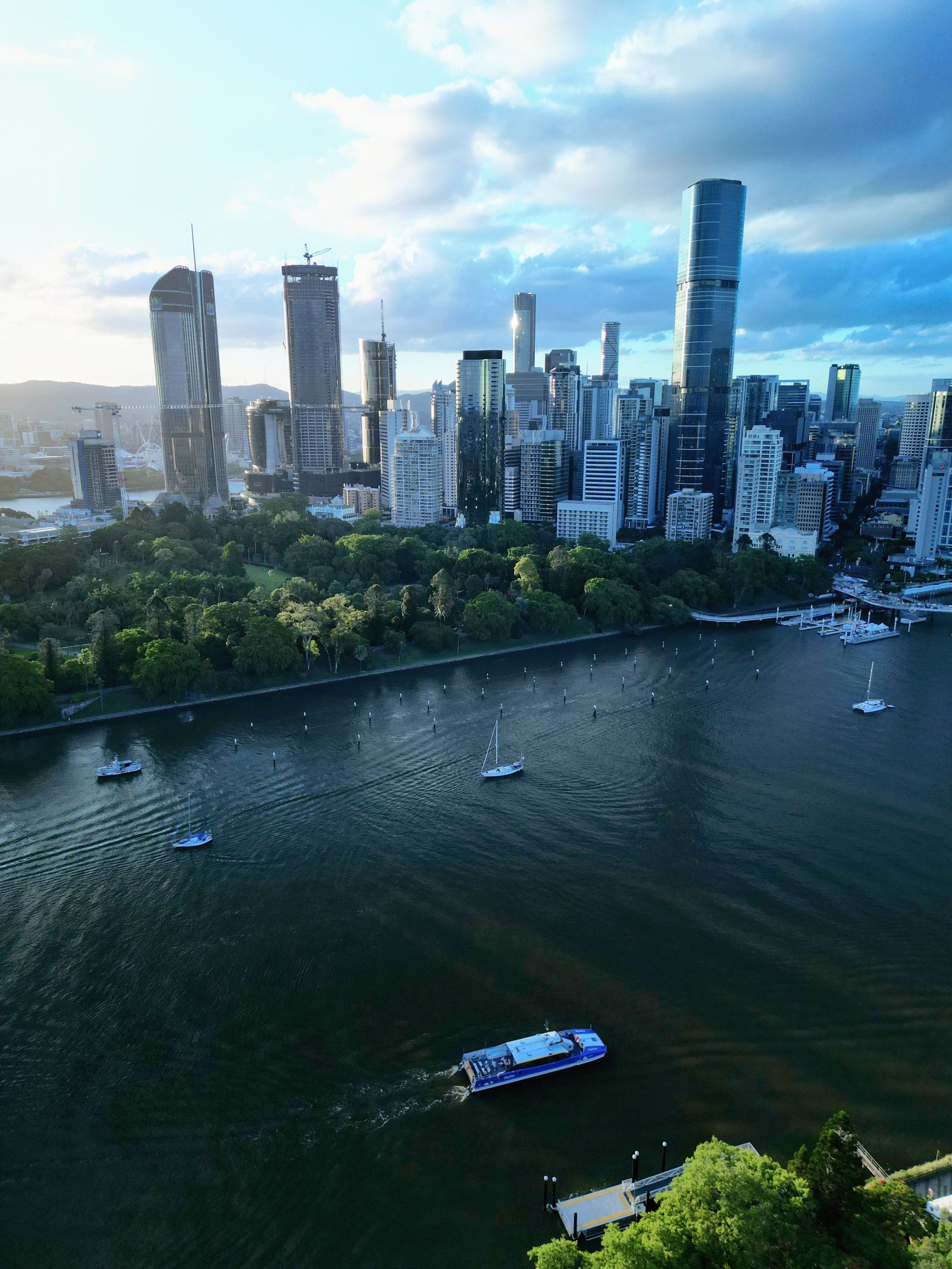 City skyline beside a river with boats and a green waterfront park under a cloudy blue sky