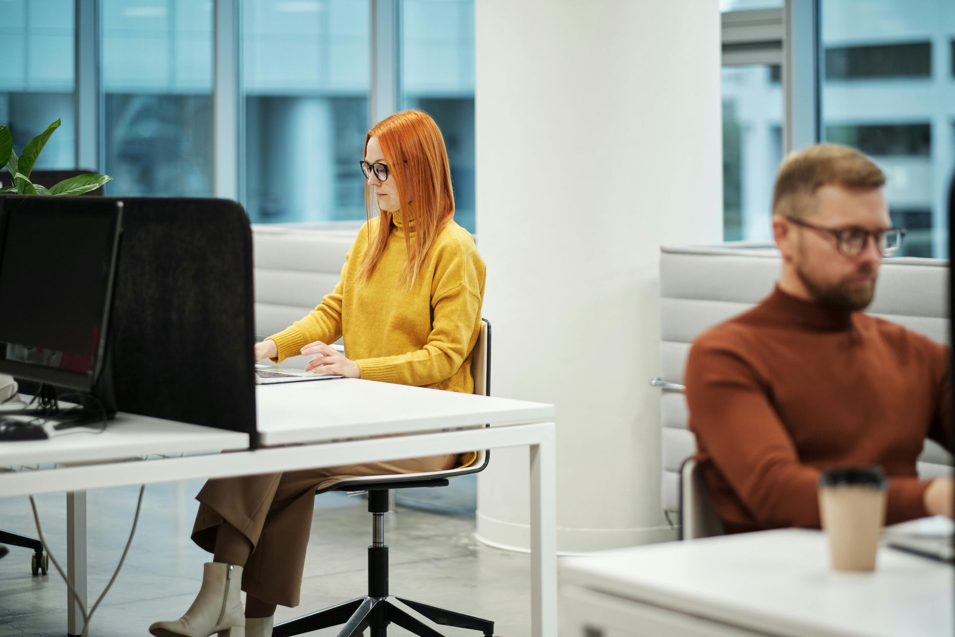 People working at desks in a bright office, with one woman in a yellow sweater at a computer.
