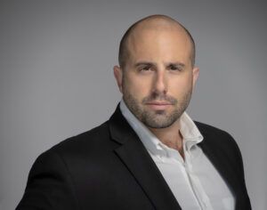 A bald man in a suit and white shirt is standing in front of a gray background.