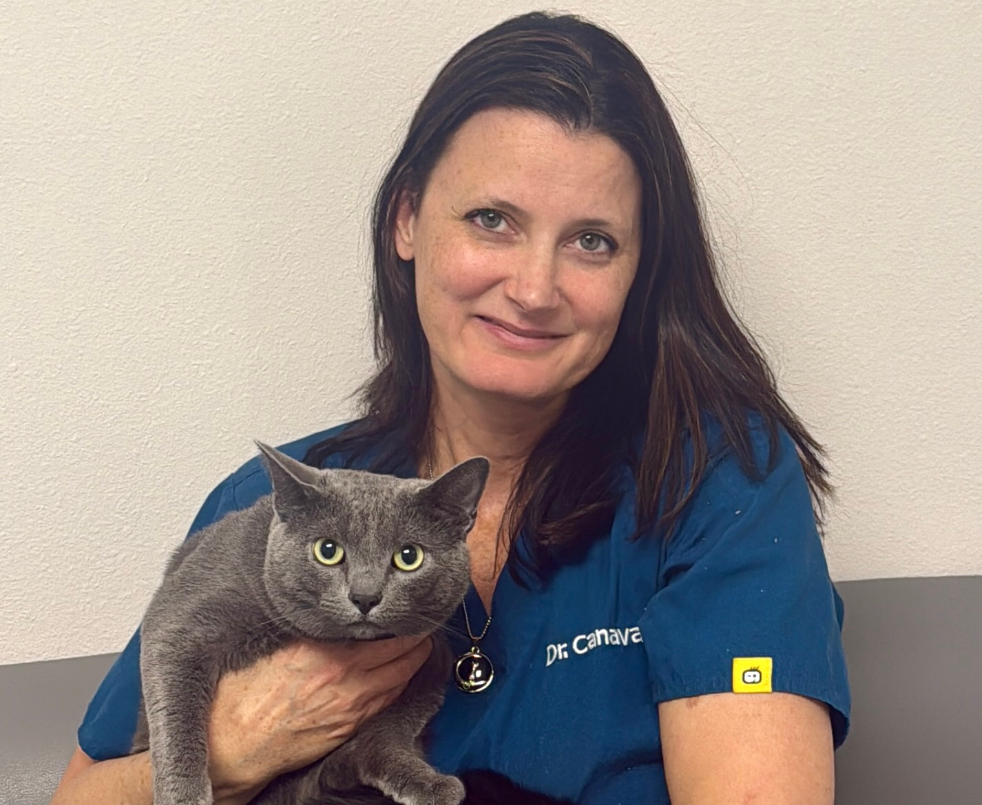 Woman in teal scrubs holds a gray and a black cat. The cats are in her lap on a gray couch.