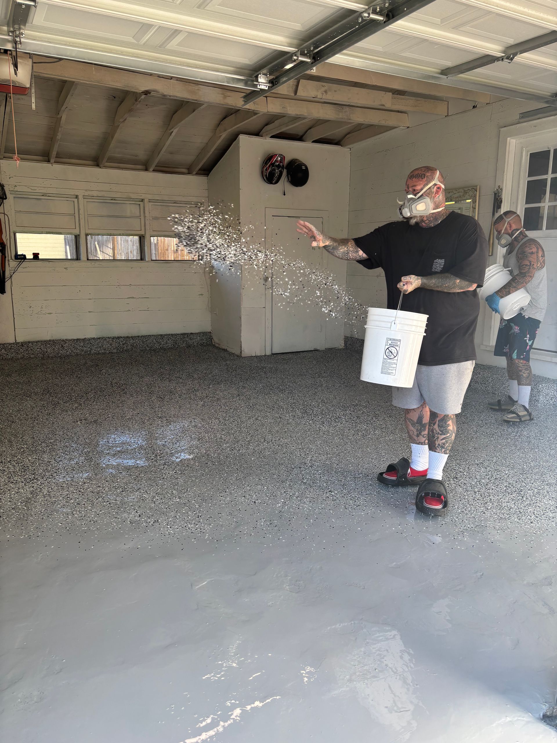Man scattering flakes on a freshly painted gray garage floor, another man in background.