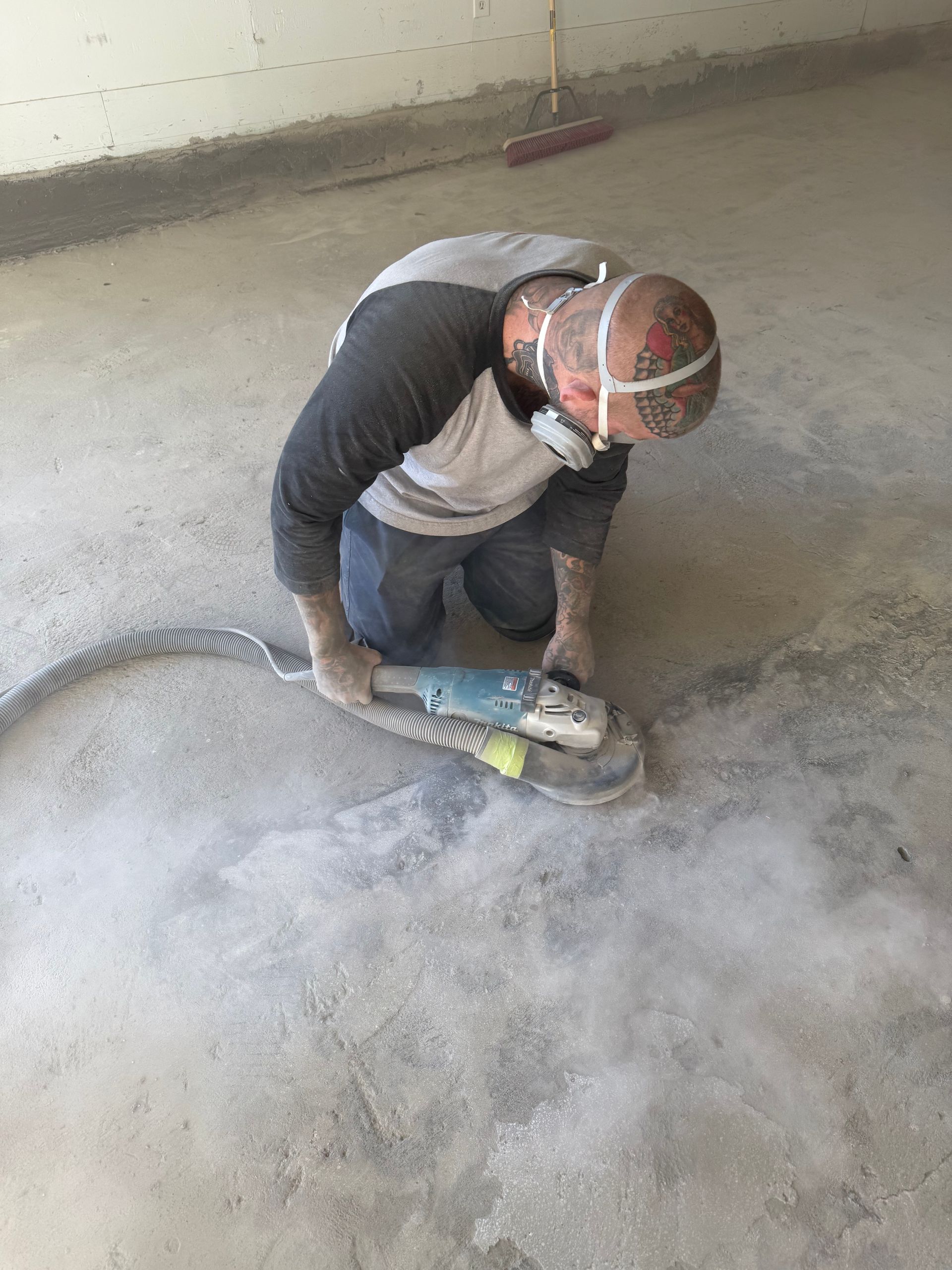 Person kneeling, grinding concrete floor, wearing respirator, generating dust.