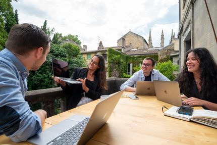 People meeting outdoors at a wooden table with laptops in a garden courtyard