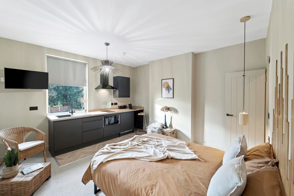 Studio apartment with bed, kitchenette, and rattan chair, in muted tones.