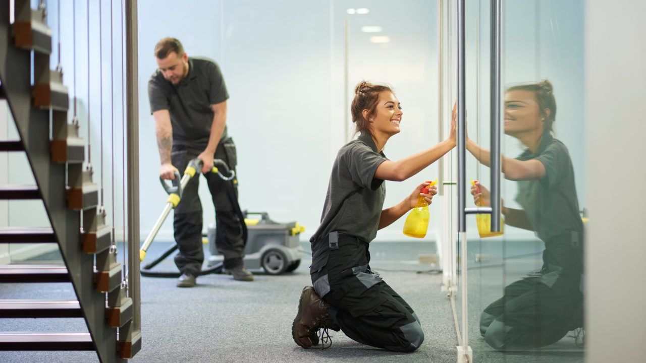 Two people cleaning an office. One vacuums, the other sprays and wipes a glass panel.