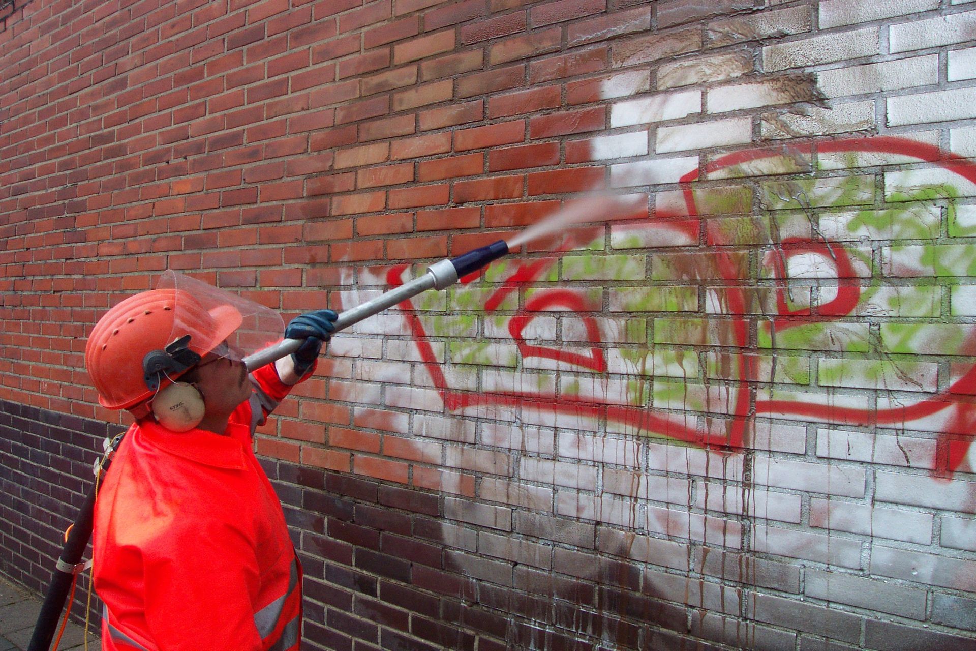 Person in orange safety gear using a pressure washer to remove graffiti from a brick wall.